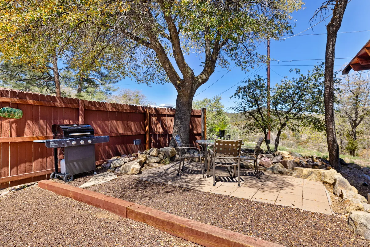 An inviting outdoor seating area surrounded by mature trees and natural rock features. A grill stands ready for use, while comfortable seating encourages gatherings. The large tree provides shade, and the open-air setting feels close to nature, suggesting an easy transition between the peaceful retreat and outdoor activities.