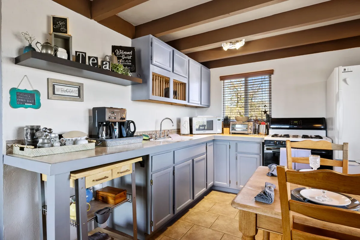 A cozy dining nook with a wooden table, coffee maker on the kitchen counter, and a window filling the space with natural light. The scene suggests intimate meals or morning coffee while enjoying views of the outdoors.